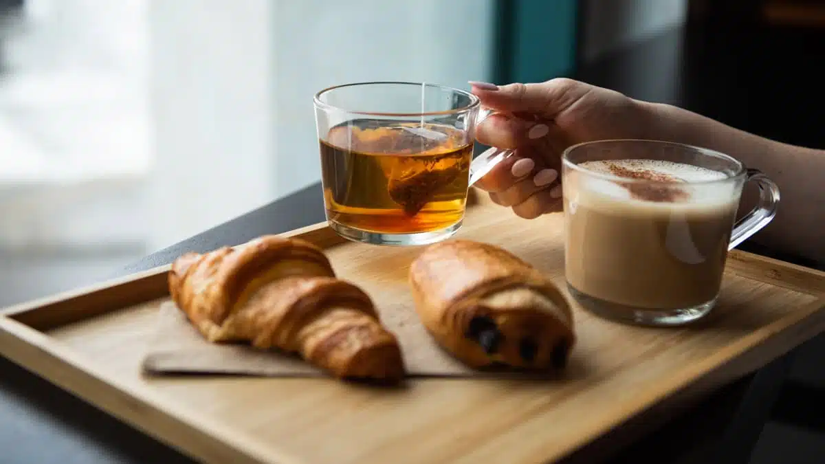 Une personne prend une tasse de thé, sur son plateau il y a des viennoiseries et un café latté