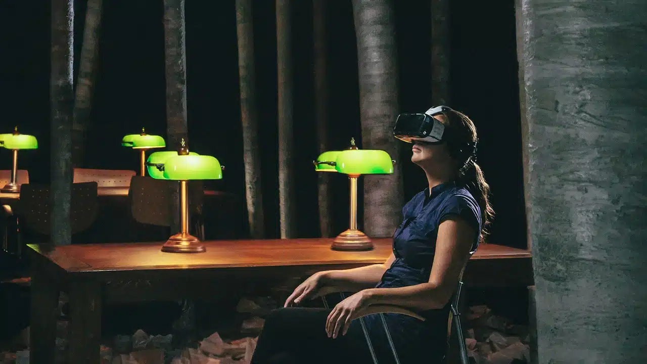 Photographie d'une femme portant des lunettes de réalité virtuelle assise à une table de bibliothèque située dans une forêt.