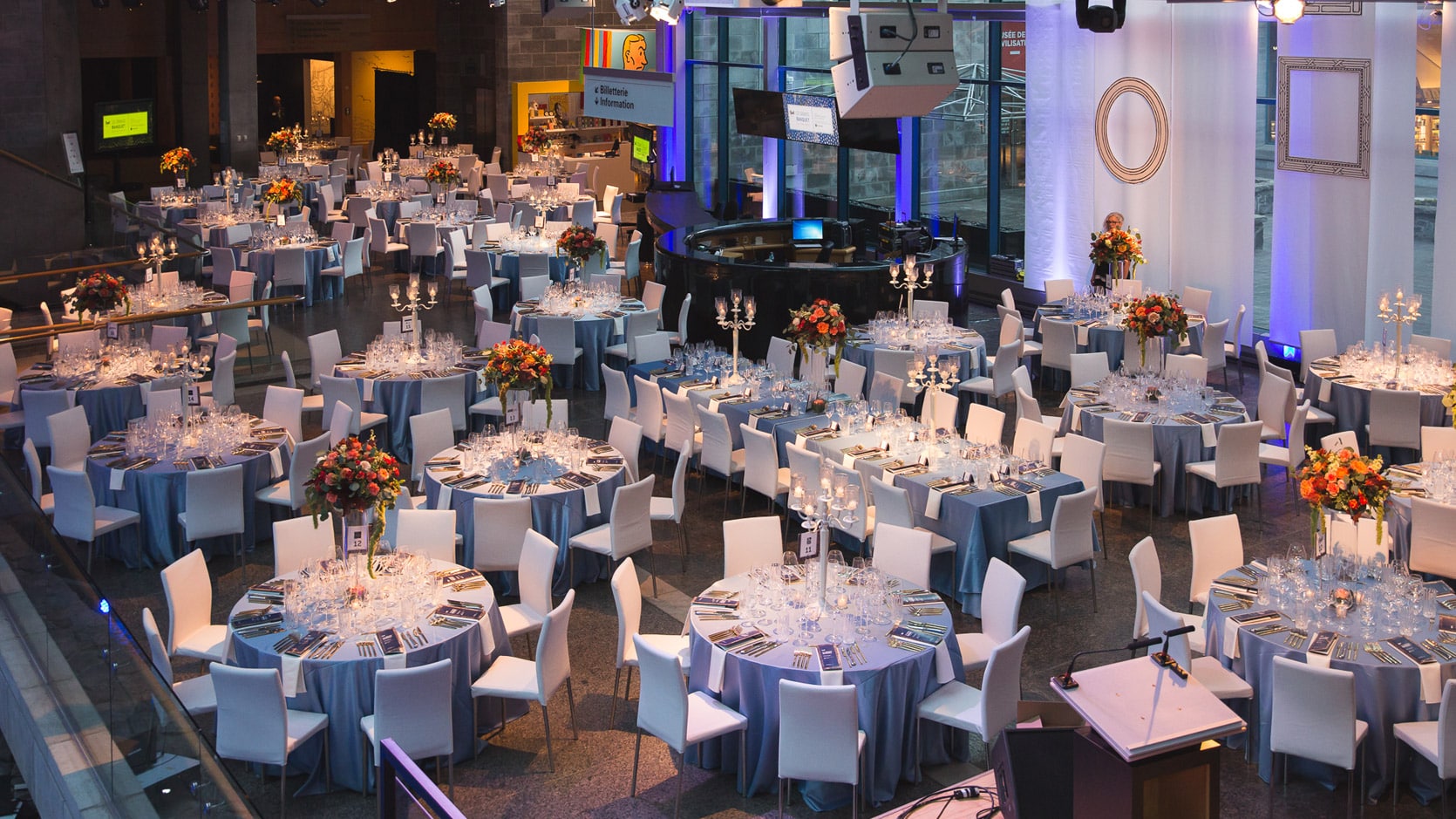 Le Hall du Musée en formule banquet, on y voit plusieurs tables rondes nappés en gris avec des chaises blanches, une grande table d'honneur et des arrangements floraux et des chandeliers.