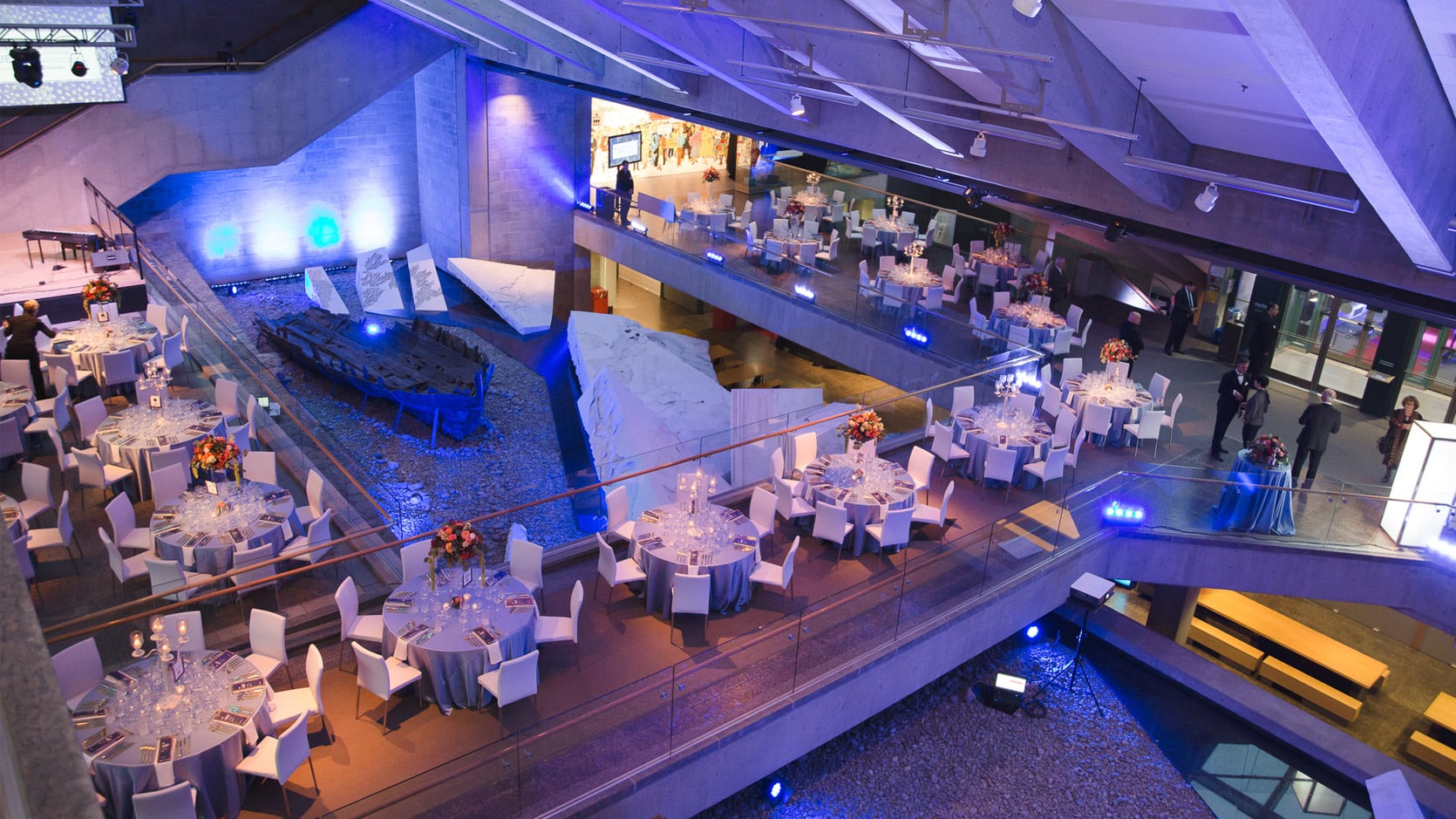 Vue en plongée du Hall du Musée en formule banquet, on y voit plusieurs tables rondes nappés en gris avec des chaises blanches et des arrangements floraux et des chandeliers.