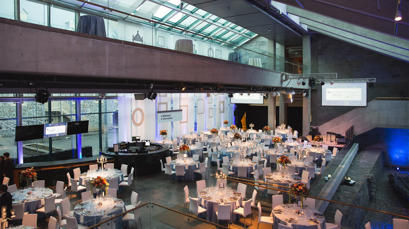 Le Hall du Musée en formule banquet, on y voit plusieurs tables rondes nappés en gris avec des chaises blanches et des arrangements floraux et des chandeliers.
