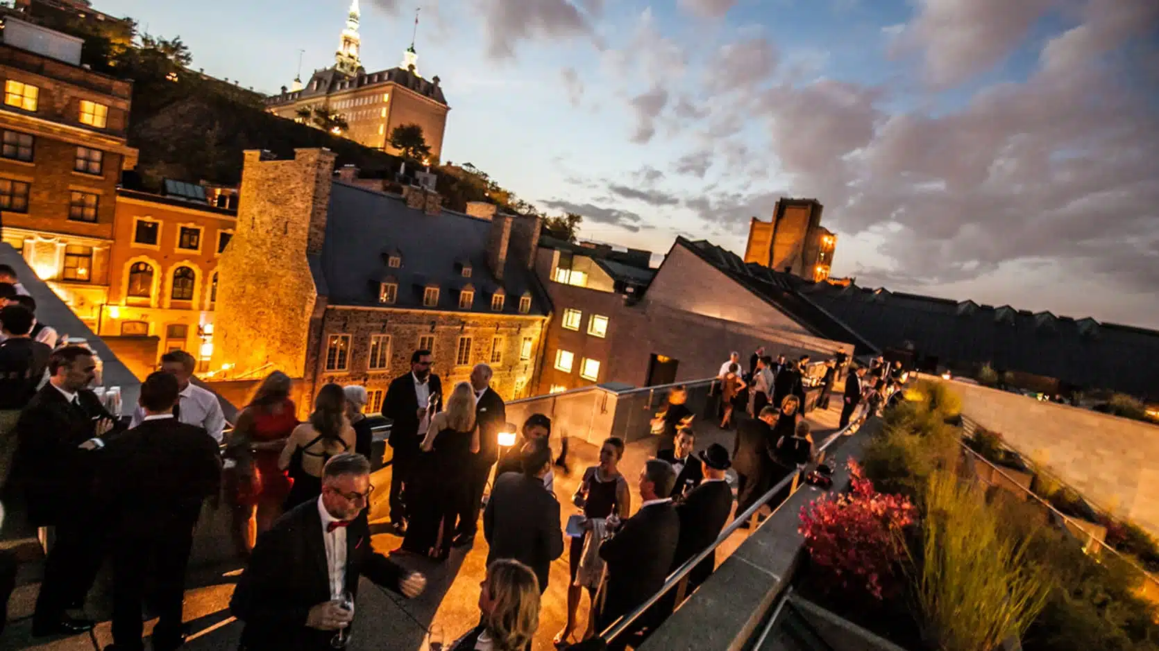 Des personnes participent à un cocktail en soirée sur les terrasses du Musée. Au fond on aperçoit le Séminaire de Québec.