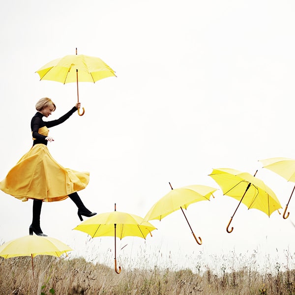 Une dame portant une robe jaune et de hautes bottes noires marche sur une envolée de parapluies jaunes en ayant un parapluie à la main.