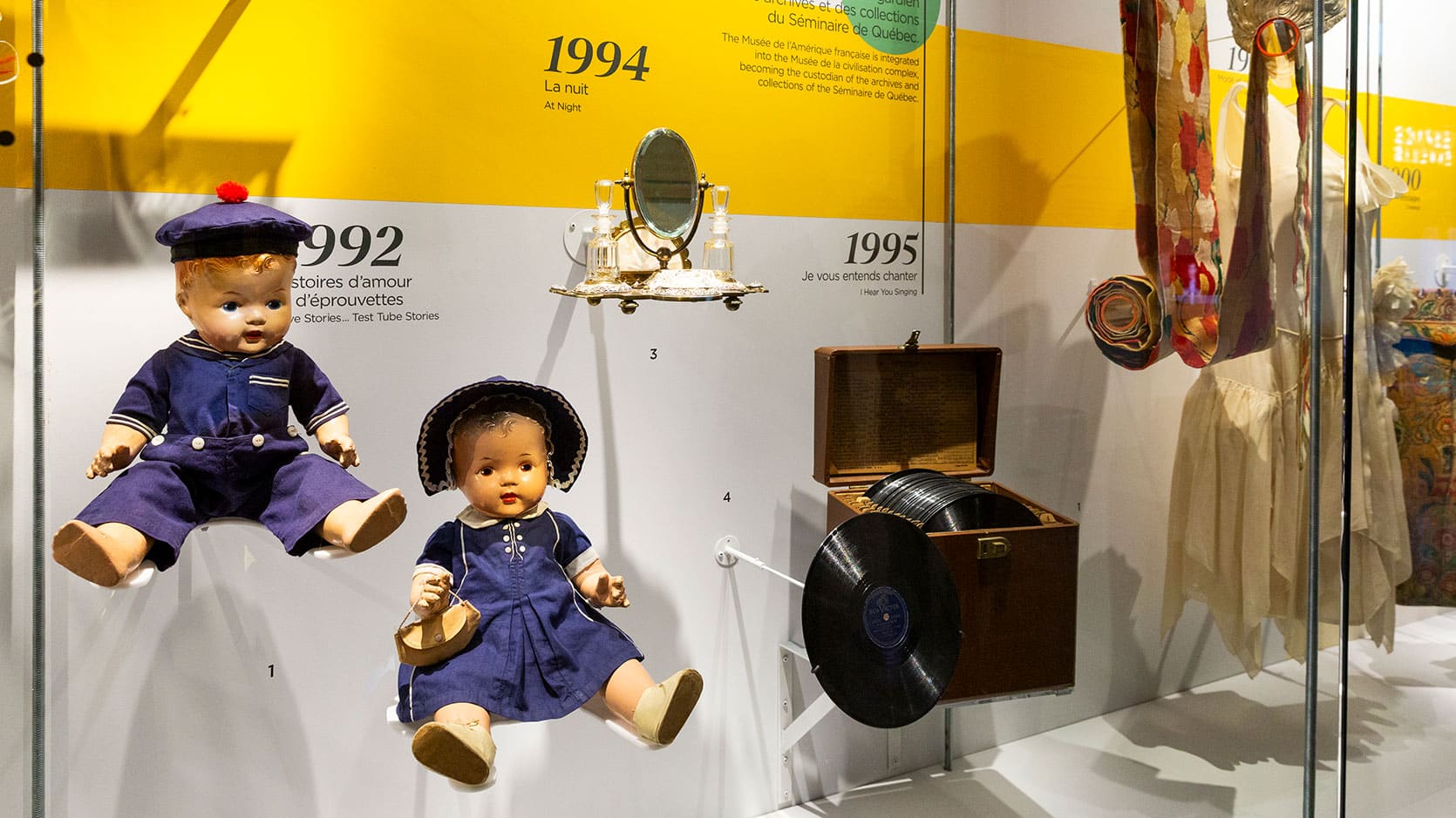Objets en vitrine alignés sur une ligne du temps représentants les expositions marquantes du Musée. On y voit des poupées, un miroir, un boîte de disques et une robe.