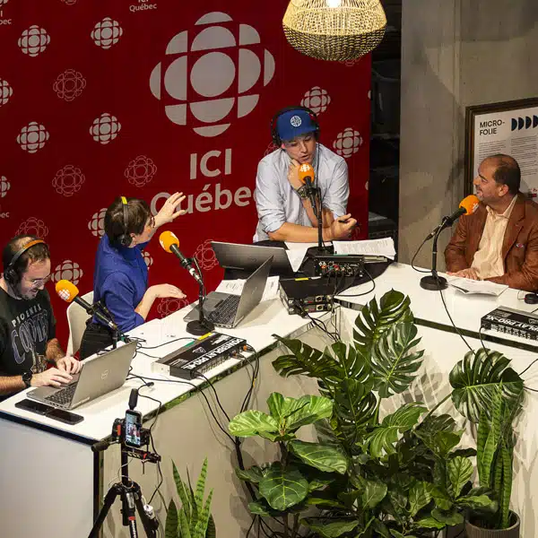 Quatre personnes sont assis sur une table blanche en angle avec un décor rouge portant le logo de Radio-Canada en blanc et la signature Ici Québec. Devant eux, ils ont des micros oranges.