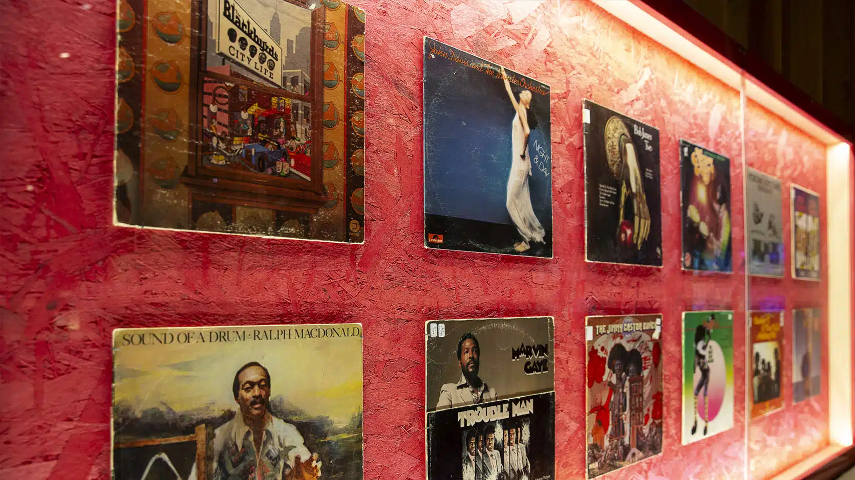 Plusieurs pochettes de disques vinyles sont accrochés dans une vitrine au fond rouge.