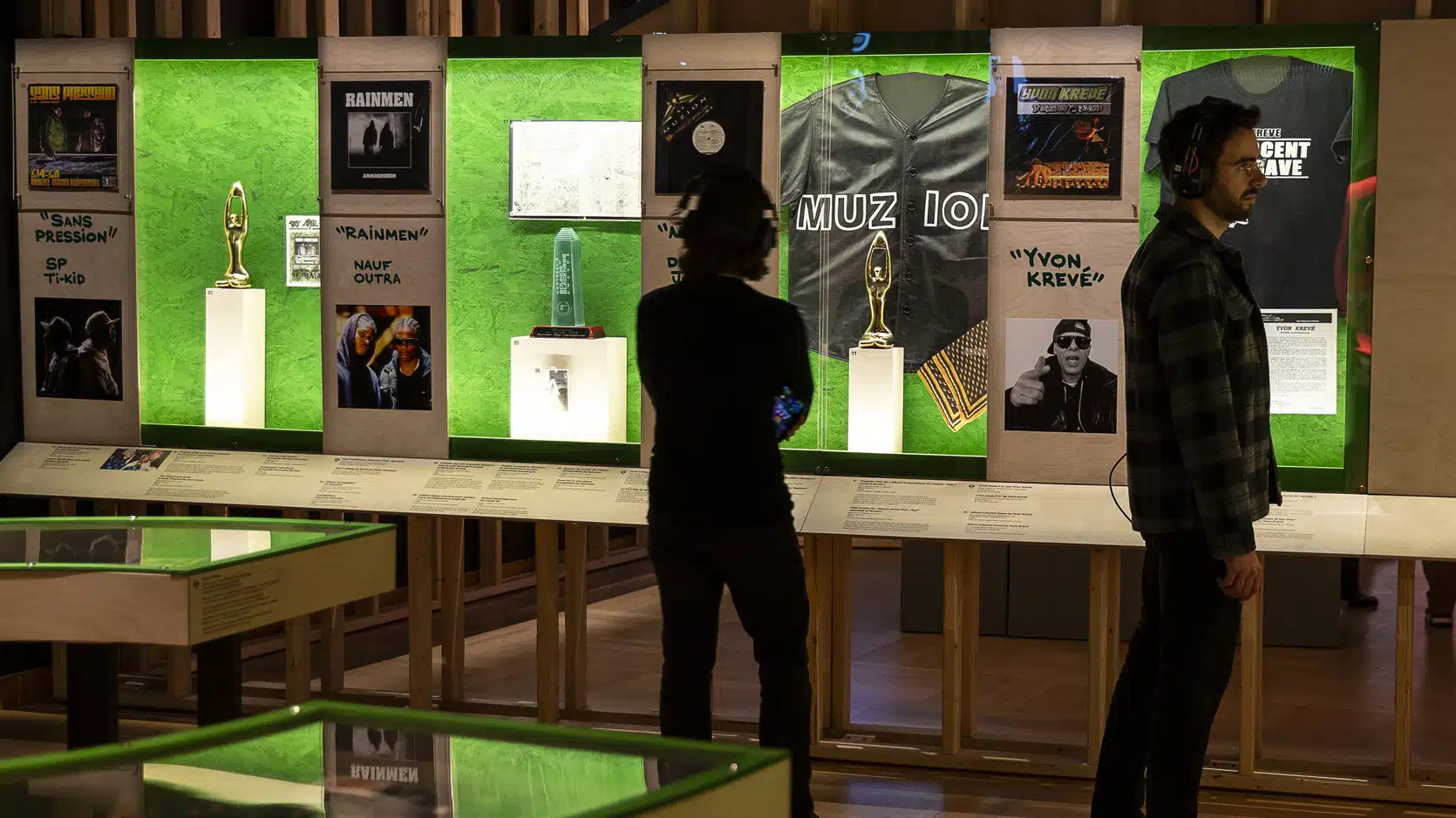 Deux personnes regardent des vitrines au fond vert dans lesquelles des prix, des vinyles et autres objets d'artisans et artisanes du hip-hop québécois sont exposés.