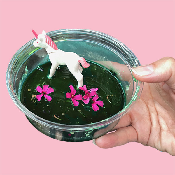 une licorne blanche à la crinière rose dans un bol en verre, rempli d'un liquide vert parsemé de fleurs roses. Le bol est tenu par une main