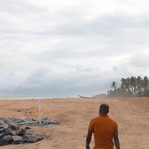 Un homme noir avec un polo orange photographié de dos sur une plage sauvage où l'on voit des pierres, des palmiers et la mer au loin
