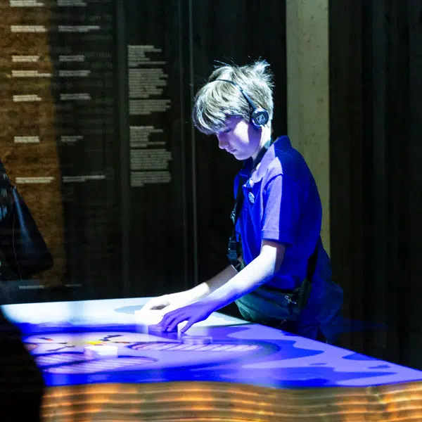 un garçon blond-châtain avec un polo manche courte bleu-mauve qui explore de la main une table où est représentée une sorte de carte