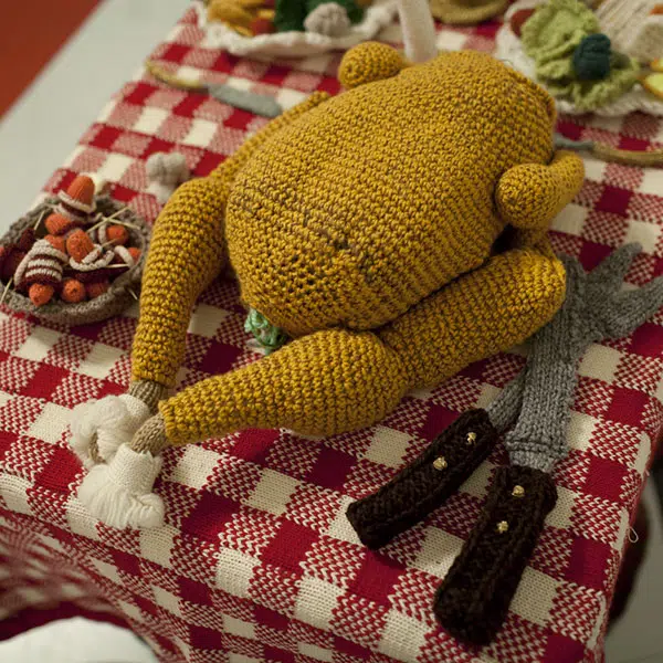 photo d'une tablée des Fêtes où l'on voit une dinde, des ustensilles et des accompagnements déposés sur une nappe à carreaux rouge et blanc le tout en tricot