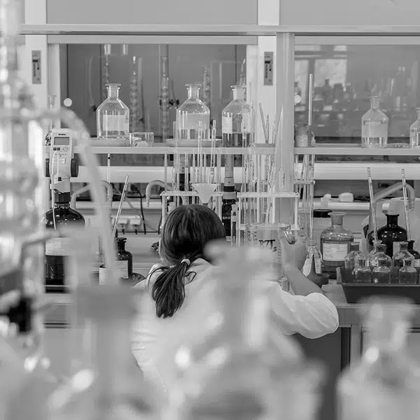 photo en noir et blanc d'un laboratoire avec des éprouvettes, bocaux où l'on voit une scientifique de dos avec un sarreau accroupie observant les résultats d'un test