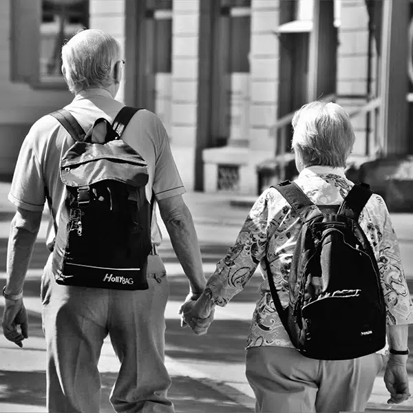 photo noir et blanc d'un couple de personnes âgées qui se tiennent la main prise de dos. L'homme et la femme portent un sac à dos