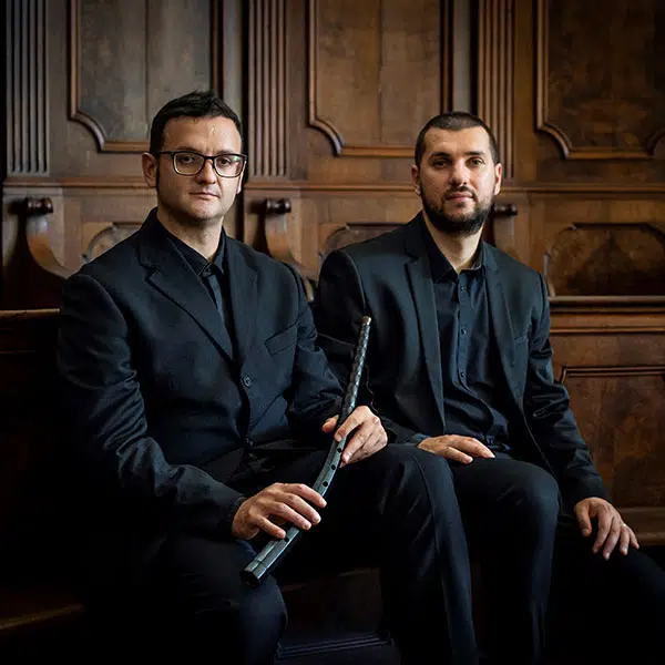 photographie d'un duo masculin, habillé en complet noir et chemise noires, assis sur un banc d'église adossé à un mur à caissons, l'un tenant une flûte de type traversière