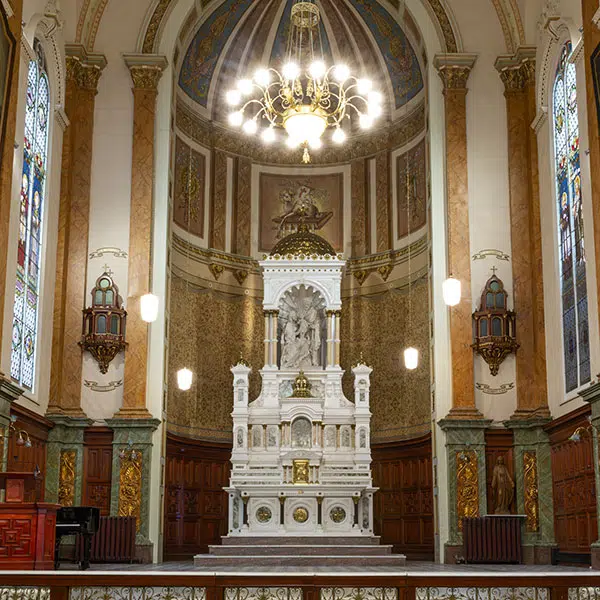 Photographie de l'autel de la Chapelle extérieure du Séminaire de Québec. L'autel est en vue frontale et est flanqué de reliquaire de part et d'autre. Un luminaire allumé est suspendu au dessus de l'autel . On peçoit une peinture de l'agneau sacrificiel sur le mur du choeur au dessus de l'autel. Dans la partie supérieure de l'autel est nichée une sculpture de la Sainte Famille. L'autel est blanc immaculé et on perçoit quelques éléments dorés.