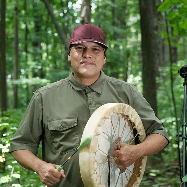 photographie d'un musicien-chanteur autochtone Shauit tenant une tambour à la main. Il porte une casquette bourgogne et une chemise kaki à manches courtes