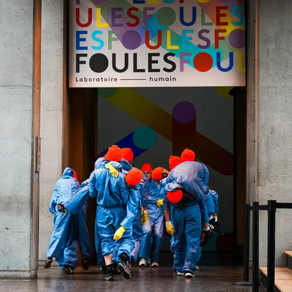 Photographie qui montrent les étudiants de l'École de cirque en pleine représentation de leur spectacle Nous sommes. Ils sont une douzaine tous de dos et rassemblés dans des positions variées. Ils sont à l'entrée de l'exposition Foules, laboratoire humain. On aperçoit une partie de l'affiche de Foules dans le haut de l'image. Les étudiants sont tous habillés avec une combinaison de travail bleue, ils portent tous un bonnet orange fluo sur la tête et des gants en caoutchouc jaune.