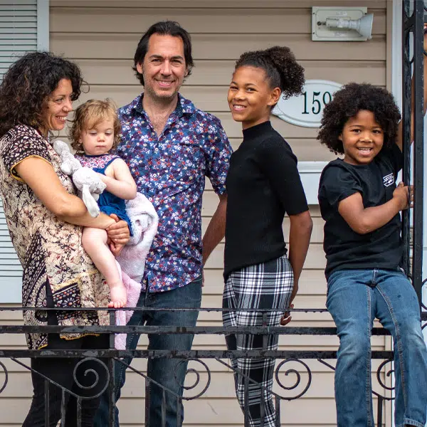 Photographie qui montre une famille de réfugiés devant l'entrée d'une maison. On voit à l'arrière plan le numéro de porte d'entrée et le mur de façade beige de la maison ainsi. La ferronnerie d'un balcon d'entrée est partiellement visible en bas et sur le côté droit de l'image. La famille est placée derrière la ferronnerie du balcon. La famille est composée, en partant de gauche à droite sur l'image, de : la mère qui tient dans ses bras une jeune fille, du père, d'une jeune adolescente et enfin d'un garçon. Le garçon est assis sur la barre supérieure du balcon et se tient à la colonne de la ferronnerie partiellemnt visible à droite.