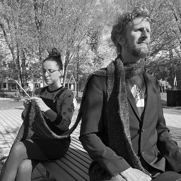 photo en noir et blanc des artistes multidisciplinaires Angèle Courville et Philippe-Emmanuel David assis sur un banc de parc, elle en train de tricoter un foulard porté par lui