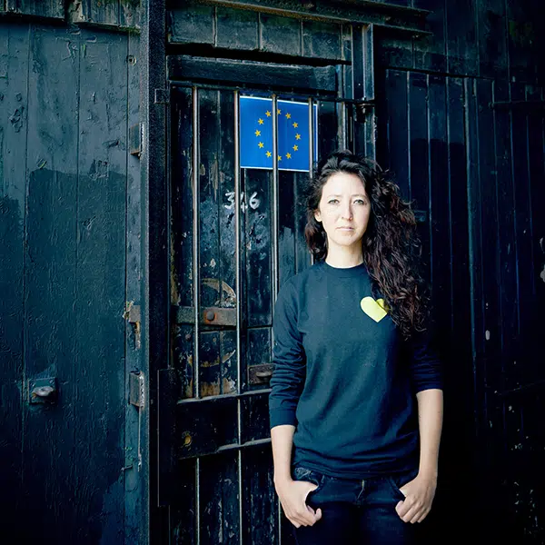 photo couleurs de l'artiste Sara Boris. Elle est adossée à un mur bleu fait de vieilles lattes de bois sur lequel est collé le drapeau de l'union européenne