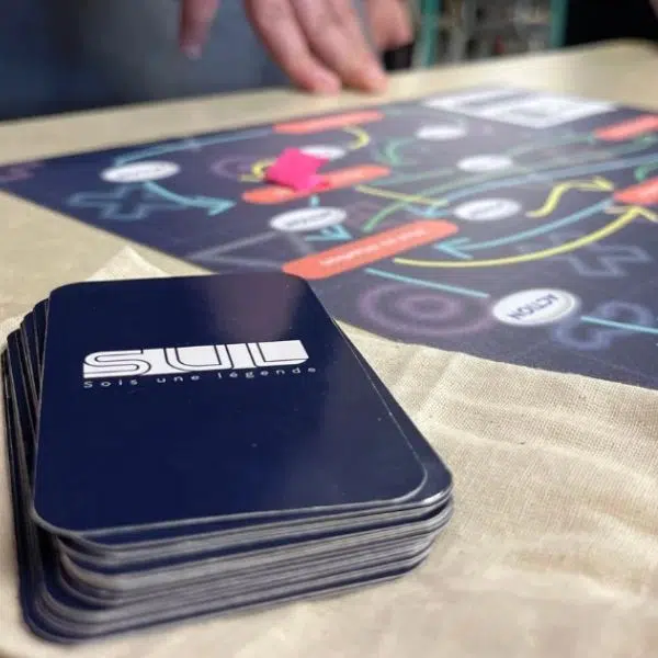 Photographie d'un jeu de cartes avec le sigle SUL qui signifie Suis une légende sur l'endos et un plateau de jeu en arrière-plan de type jeu de société