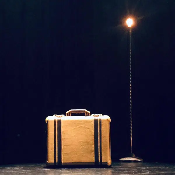 une valise beige avec des lanières noires déposée sur une scène à côté d'une lampe sur pied sans abat-jour