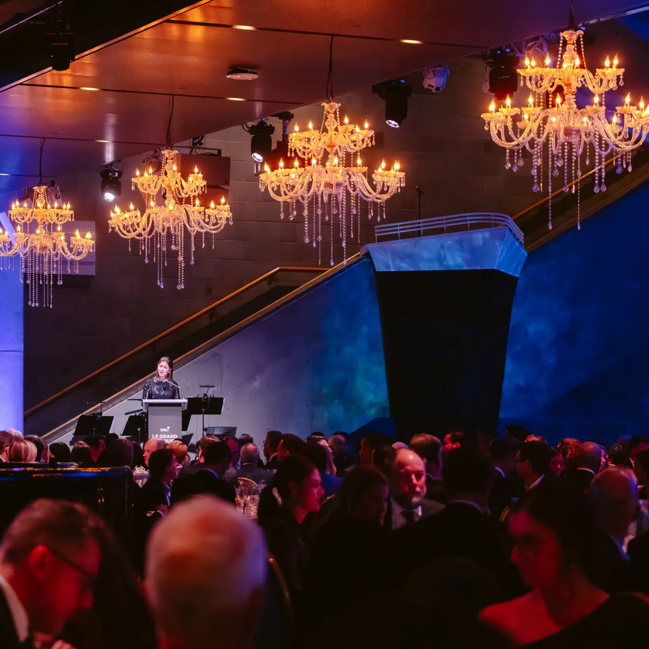 Dans le Hall du Musée plusieurs personnes sont assises devant une oratrice. Au plafond de gigantesques lustres de style chandeliers et au fond une coque de bateau.