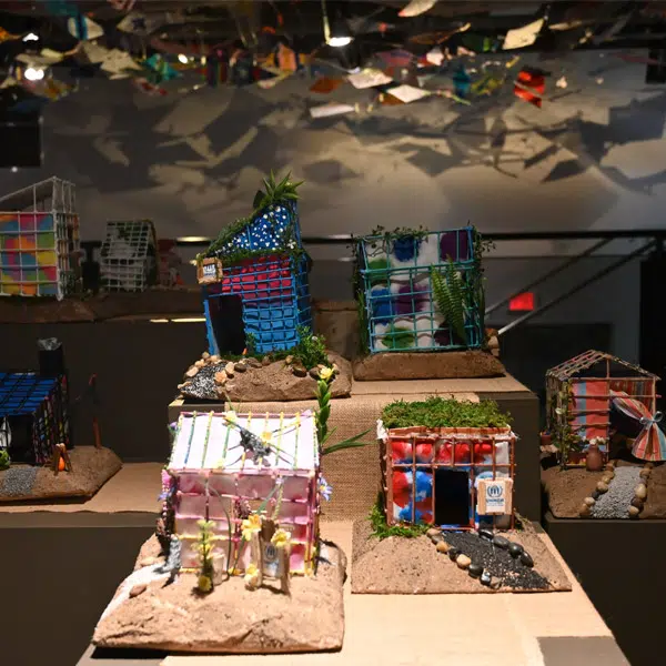 Photographie prise dans l'exposition Sous le même ciel, dans Voie libre. Elle montre des maisons de réfugiés en avant plan et des cerfs-volants en arrière plan.