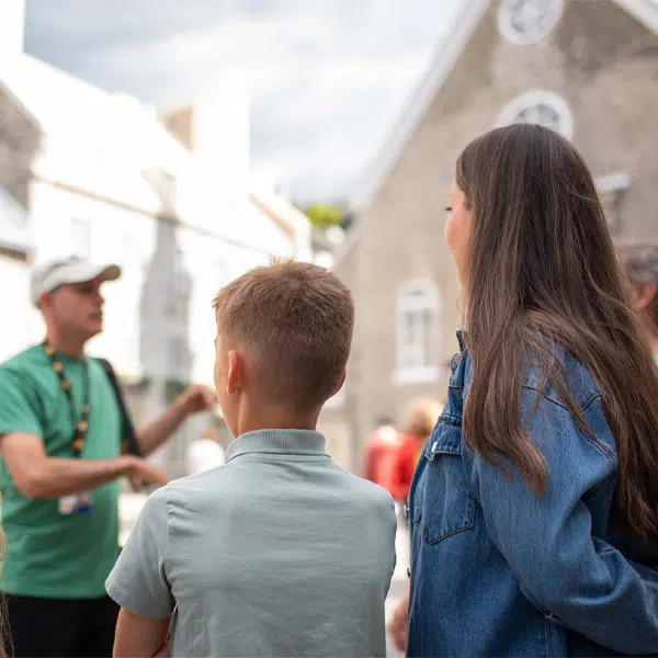 Photo prise sur la Place Royale à Québec. On voit en avant plan deux adolescents de dos. à l'arrière on reconnaît en flou un guide du musée et l'Église Notre-Dame-des-Vistoires
