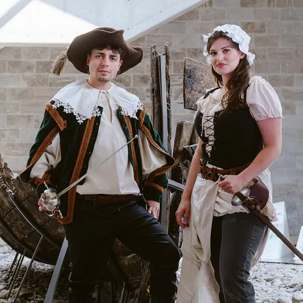 Un homme et une femme habillés en vêtements de la Nouvelle-France posent devant la barque dans le Hall du Musée